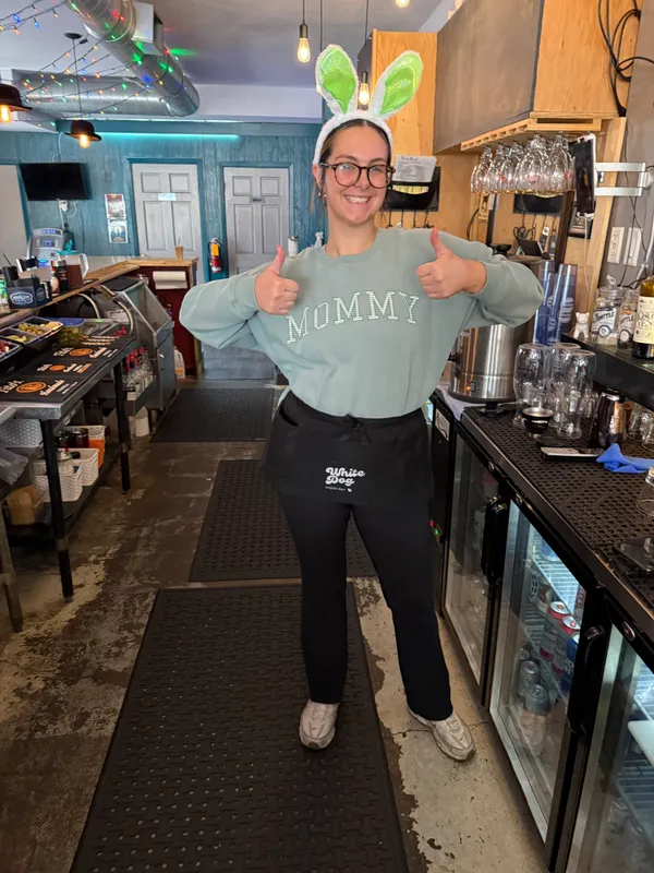 A White Dog staff member in Easter bunny ears and apron giving two thumbs up behind the bar