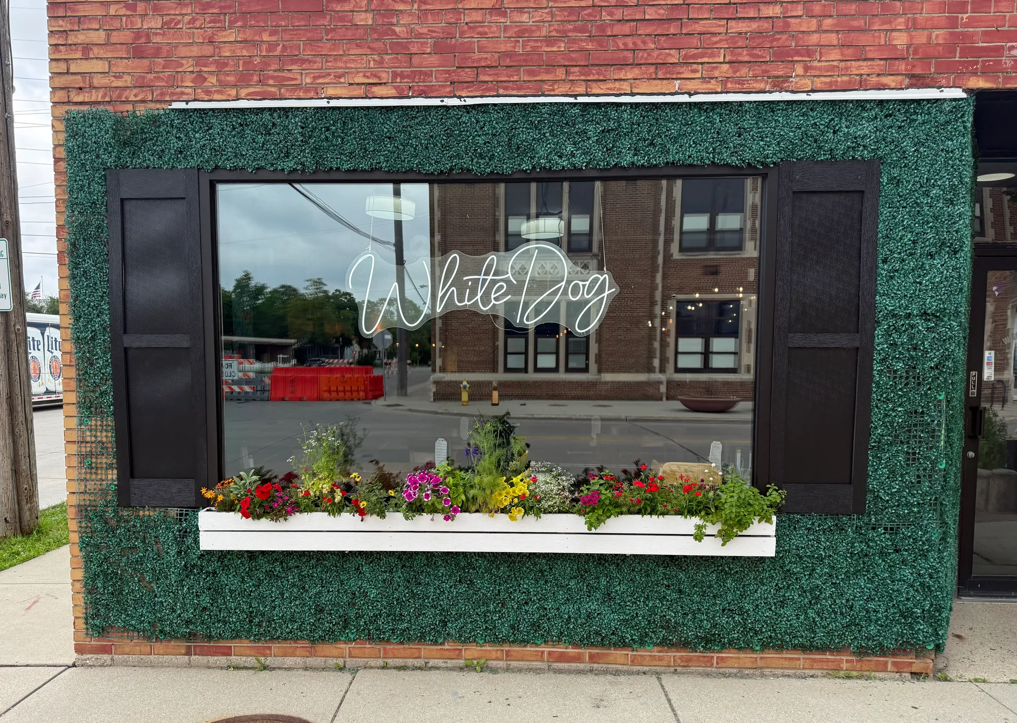 The White Dog storefront window with its neon script sign, framed by green hedging and a flower box of colorful blooms
